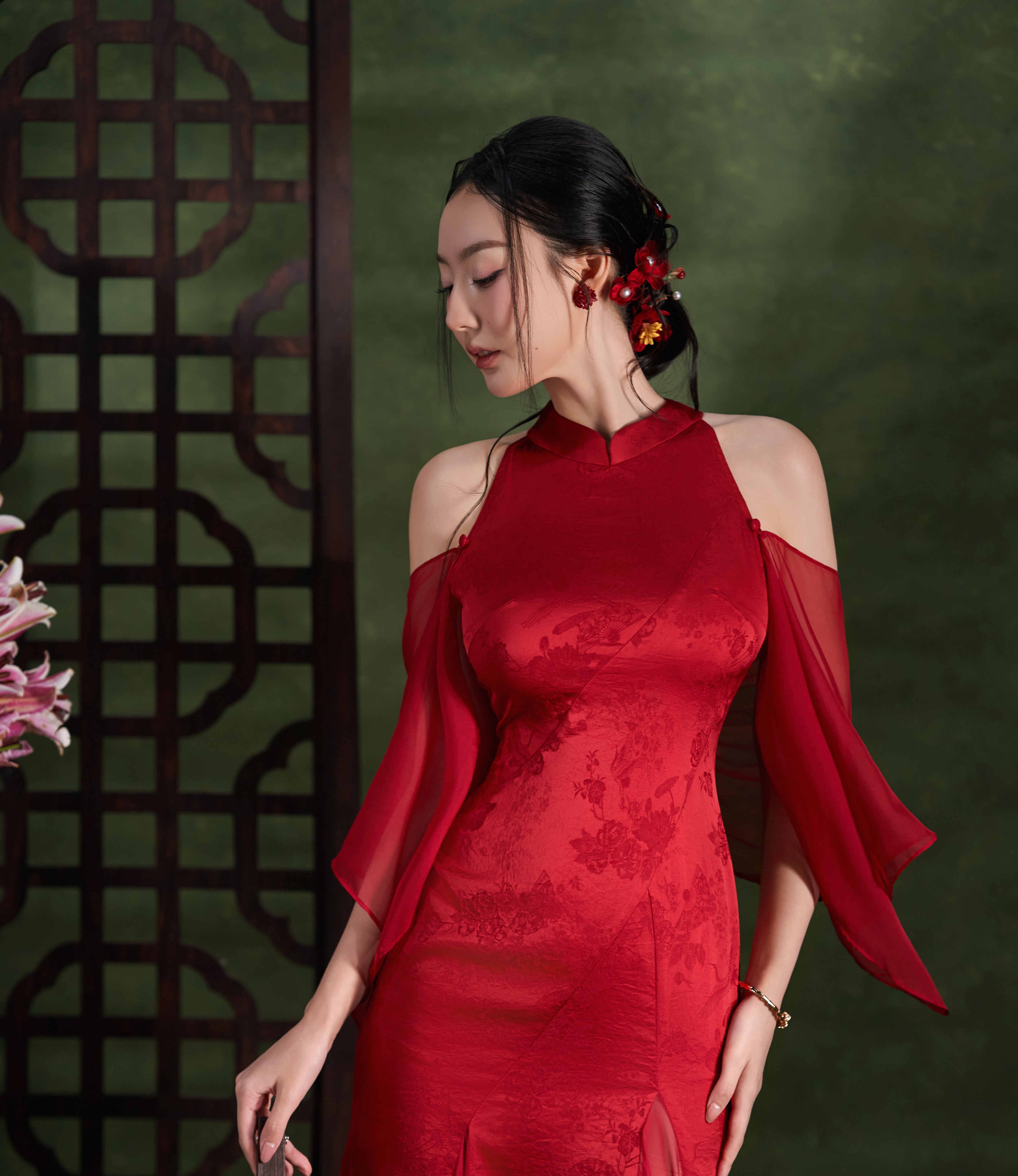 A woman wearing a red off-the-shoulder qipao dress with sheer flutter sleeves and floral embroidery, standing in front of a dark green background with a decorative screen.