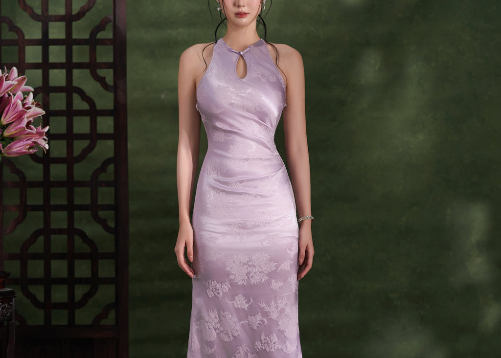 A woman stands in a studio wearing a sleeveless lavender-purple cheongsam-style dress with a floral gradient pattern that fades from light at the shoulders to darker purple at the hem. She is posed in front of a dark green textured backdrop with a dark wooden screen and pink flowers on the left.