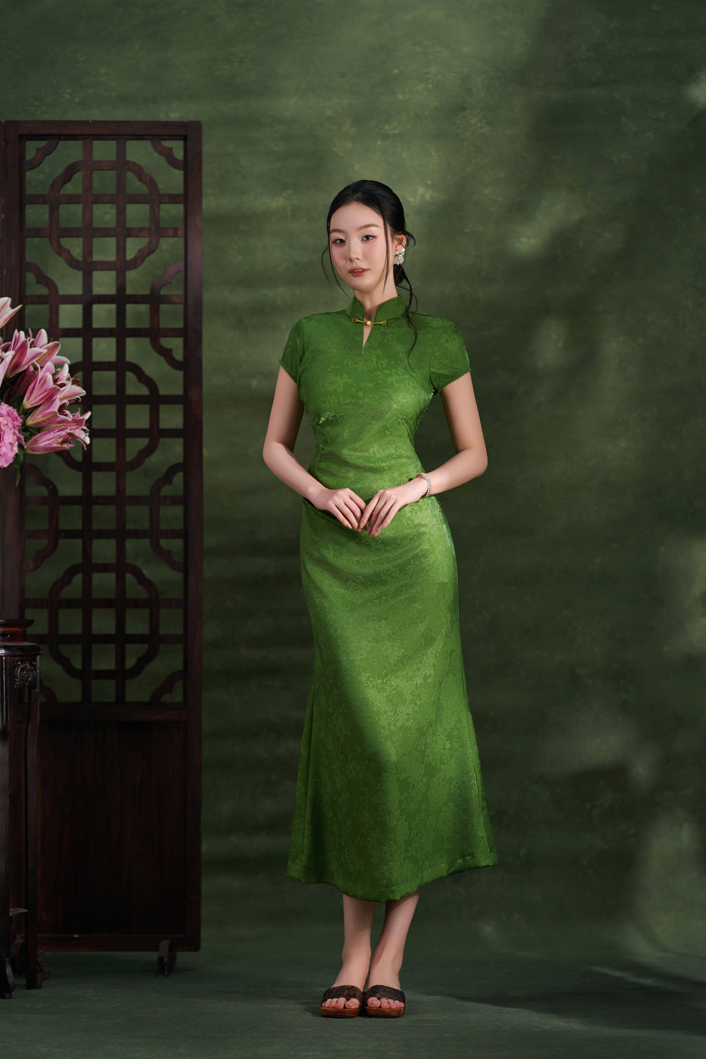 A woman wearing a green short-sleeve cheongsam dress stands in front of a textured green backdrop, with a dark wooden screen and pink flowers visible on the left.