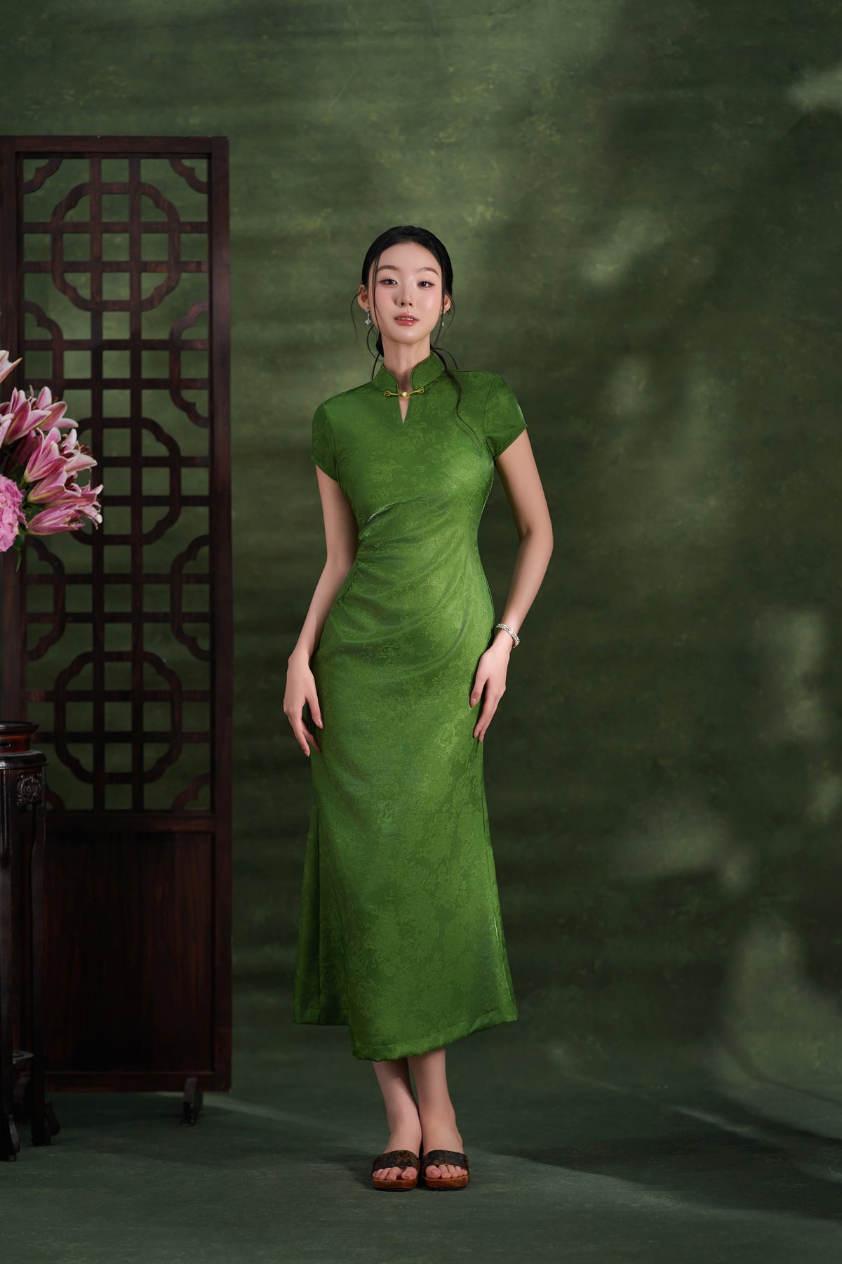 A woman stands in a studio wearing a fitted sage green cheongsam-style dress with short sleeves and a high collar. The background is a textured dark green wall with soft shadow effects, a dark wooden screen on the left, and pink flowers at the lower left edge.