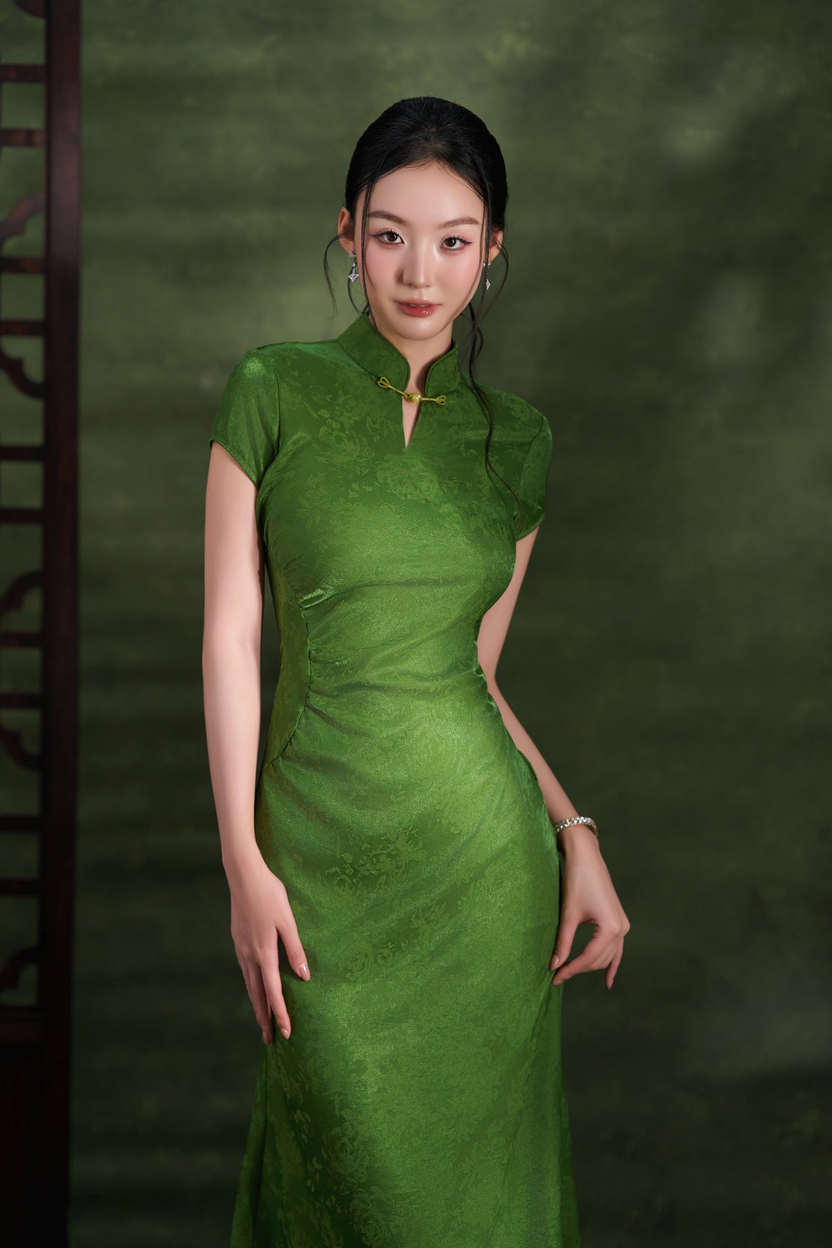 A woman wearing a green short-sleeve cheongsam-style dress with a high collar and gold button detail, standing against a dark green textured backdrop.