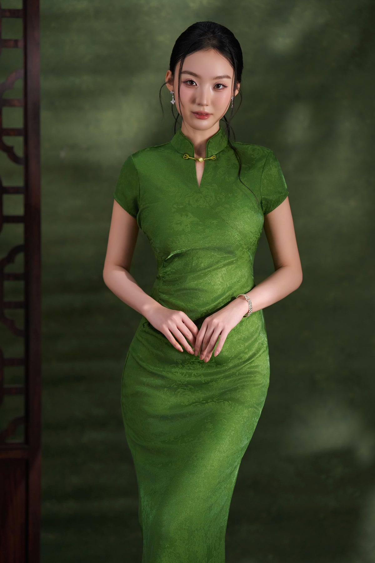 A woman wearing a fitted green short-sleeve cheongsam-style dress with a high collar and front slit, standing against a dark green textured backdrop.