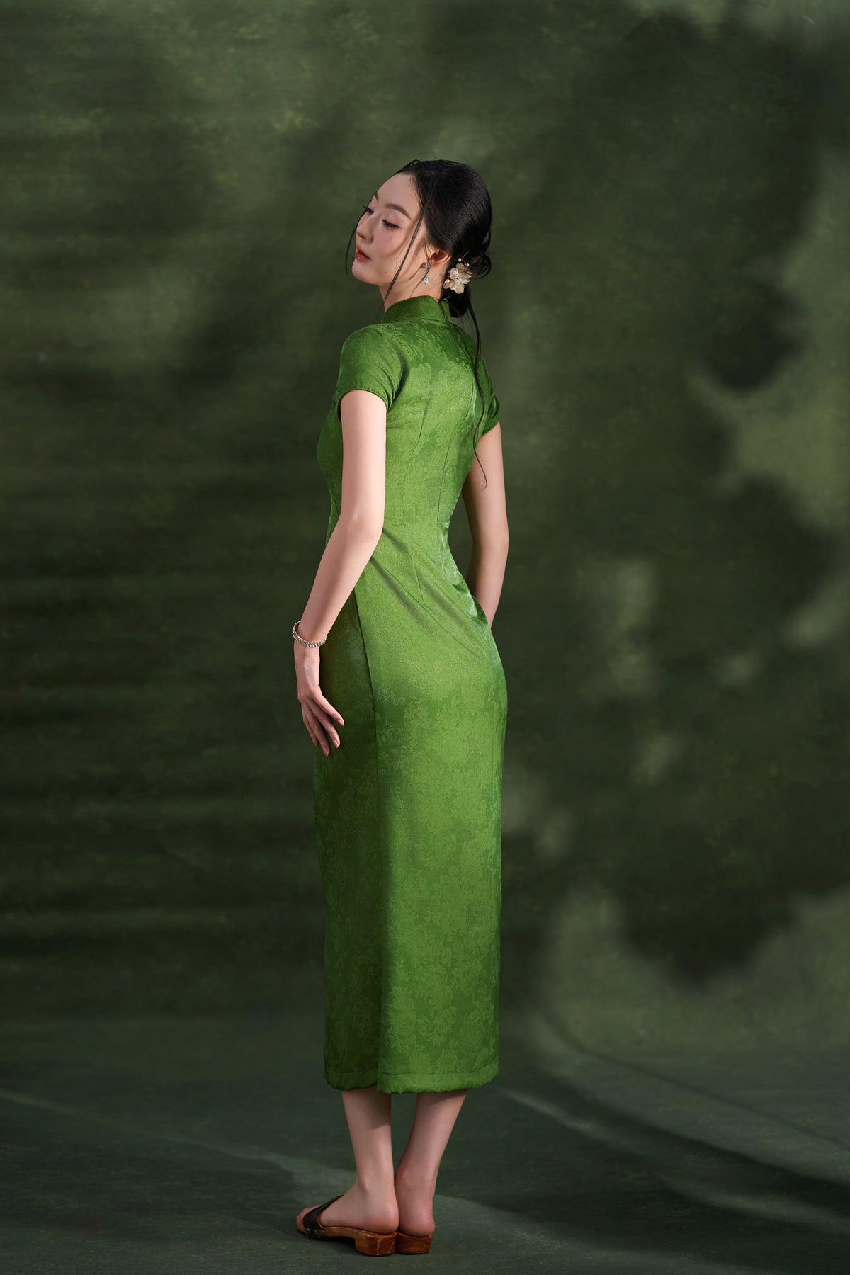 A woman wearing a green short-sleeve cheongsam dress, photographed from the back against a dark green textured studio backdrop.